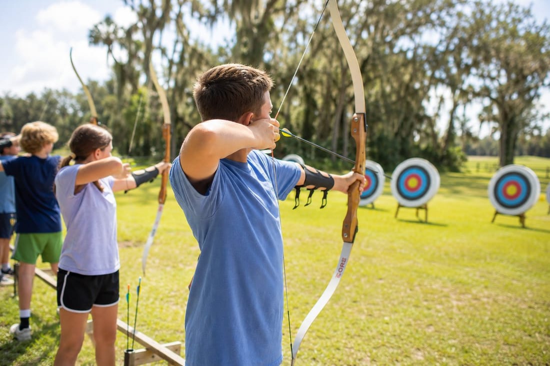 A camper drawing a bow at the archery range, learning proper form and target focus